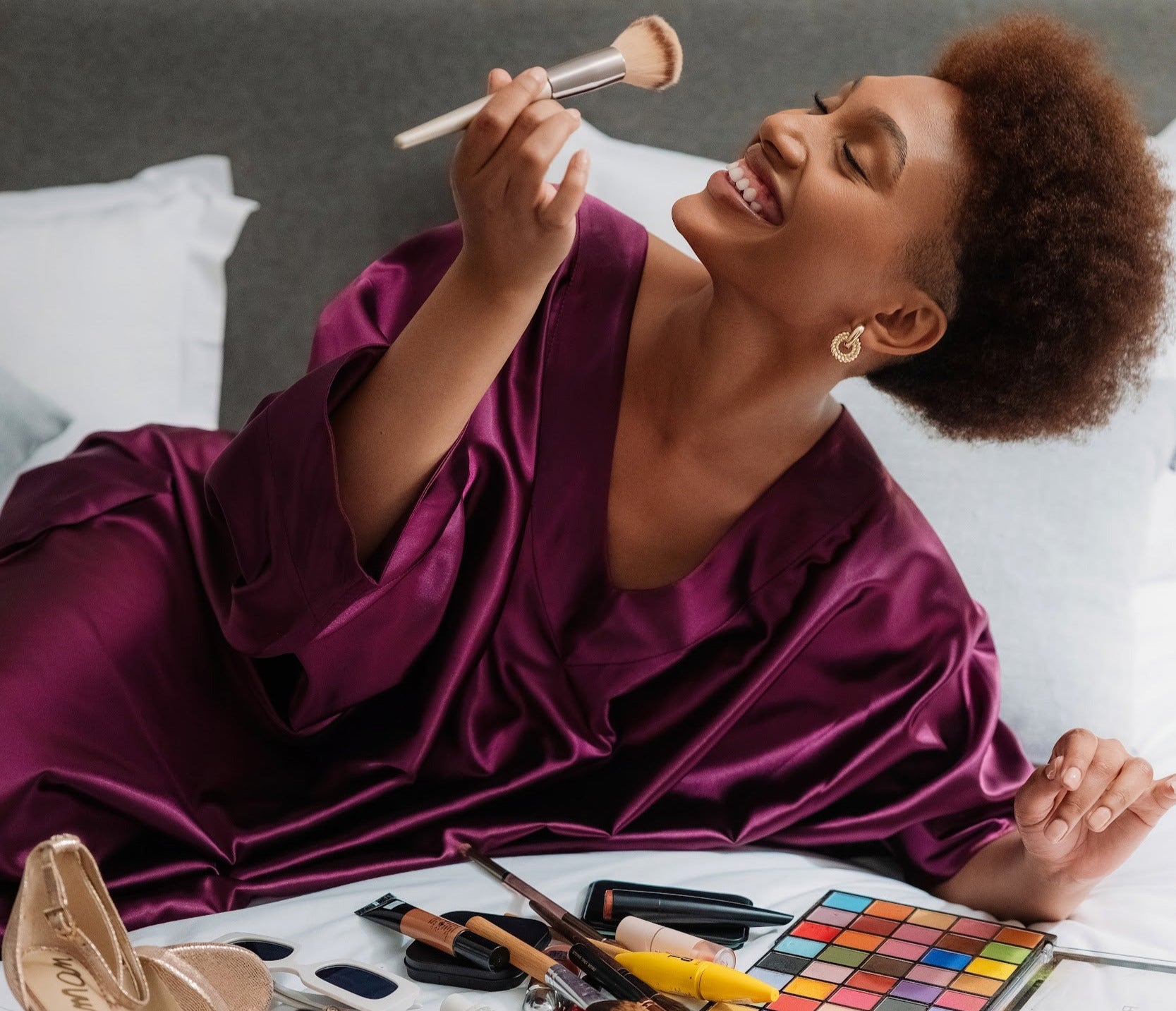 Woman applying makeup with a brush on a bed surrounded by makeup products